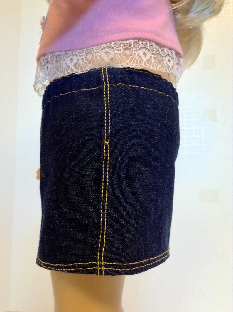 K&ouml;nnte beinhalten: Ein dunkelblauer Jeansrock mit gelben N&auml;hten. Der Rock ist Teil eines Puppenoutfits und wird mit einem rosa Oberteil mit wei&szlig;er Spitzenborte kombiniert. Der Rock hat ein klassisches, schlichtes Design.