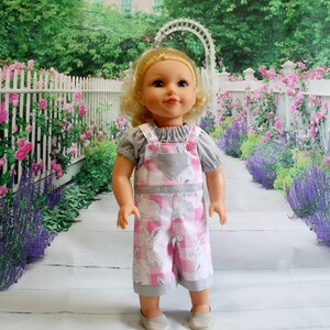 May include: A doll wearing a pink and white checkered overall with a bunny print. The overall has a gray pocket and pink buttons. The doll has blonde hair and blue eyes.
