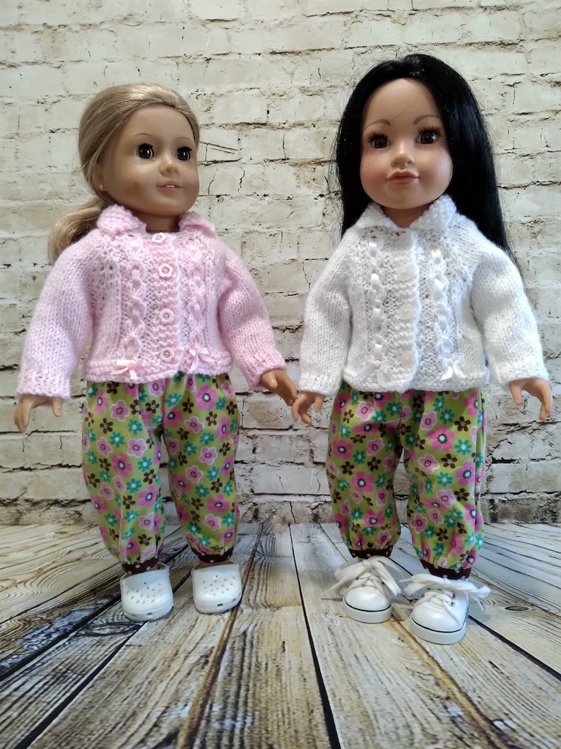 Puede incluir: Dos mu&ntilde;ecas con pantalones a juego de estampado floral y c&aacute;rdigans de punto. Una mu&ntilde;eca lleva un c&aacute;rdigan rosa y la otra un c&aacute;rdigan blanco.