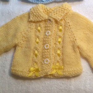 Puede incluir: Un c&aacute;rdigan de punto amarillo para beb&eacute; con cierre de bot&oacute;n blanco y detalles de cinta amarilla.