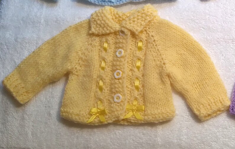 Puede incluir: Un c&aacute;rdigan de punto amarillo para beb&eacute; con cierre de bot&oacute;n blanco y detalles de cinta amarilla.