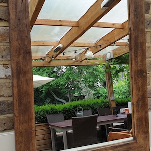 May include: A rustic wooden framed mirror reflecting an outdoor patio with a wooden pergola, string lights, and a table with chairs.