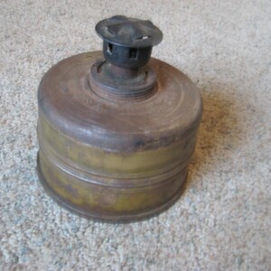 May include: A vintage, rusty, metal gas mask filter with a black metal top. The filter is a cylindrical shape with a ribbed design and a greenish-brown color.