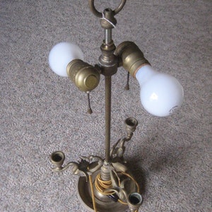 May include: A brass table lamp with three light sockets and a decorative base. The lamp has a ring at the top for carrying.