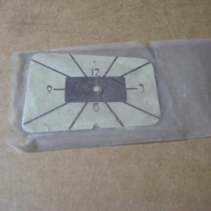 May include: A rectangular, off-white card with black lines and numbers 3, 6, 9, and 12, likely a tool or template. The card is inside a clear plastic sleeve, resting on a tan surface.