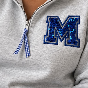 May include: A light gray quarter-zip sweatshirt featuring a large letter M patch on the left chest. The letter is filled with blue sequins and outlined in gray. A blue and white gingham ribbon is attached to the silver metal zipper pull.