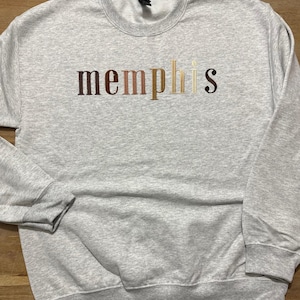 May include: A light gray sweatshirt with the embroidered word "memphis" across the chest. The lettering is in a gradient of brown, tan, and gold colors. The sweatshirt has a crew neck and long sleeves.