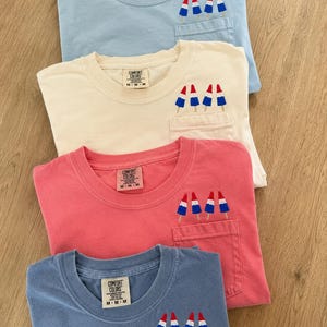 Op de afbeelding: Vier T-shirts met korte mouwen in lichtblauw, cr&egrave;me, koraal en leiblauw, elk voorzien van een borstzak met borduursel van drie rood-wit-blauwe ijsjes. Elk shirt heeft een Comfort Colors merklabel aan de binnenkant van de halslijn.