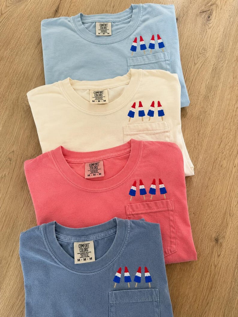 Op de afbeelding: Vier T-shirts met korte mouwen in lichtblauw, cr&egrave;me, koraal en leiblauw, elk voorzien van een borstzak met borduursel van drie rood-wit-blauwe ijsjes. Elk shirt heeft een Comfort Colors merklabel aan de binnenkant van de halslijn.