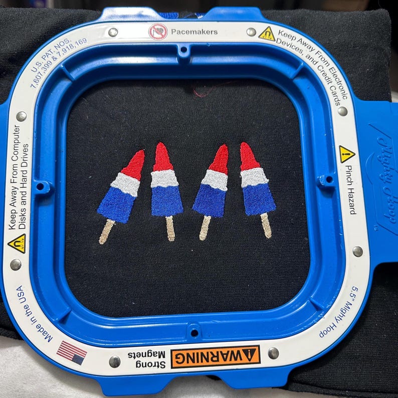 Op de afbeelding: Een blauwe 5,5 inch Mighty Hoop borduurring met zwarte stof waarop vier rood, wit en blauwe ijsjes zijn geborduurd. De ring bevat waarschuwingsteksten over magneten, pacemakers en elektronische apparaten in het Engels.