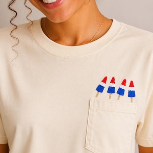 Op de afbeelding: Een cr&egrave;mekleurig T-shirt met korte mouwen en een borstzak. Boven de zak zijn vier kleine ijsjes geborduurd, elk met een rode bovenkant, een witte middenkant en een blauwe onderkant, afgewerkt met een lichtbruin houten stokje.