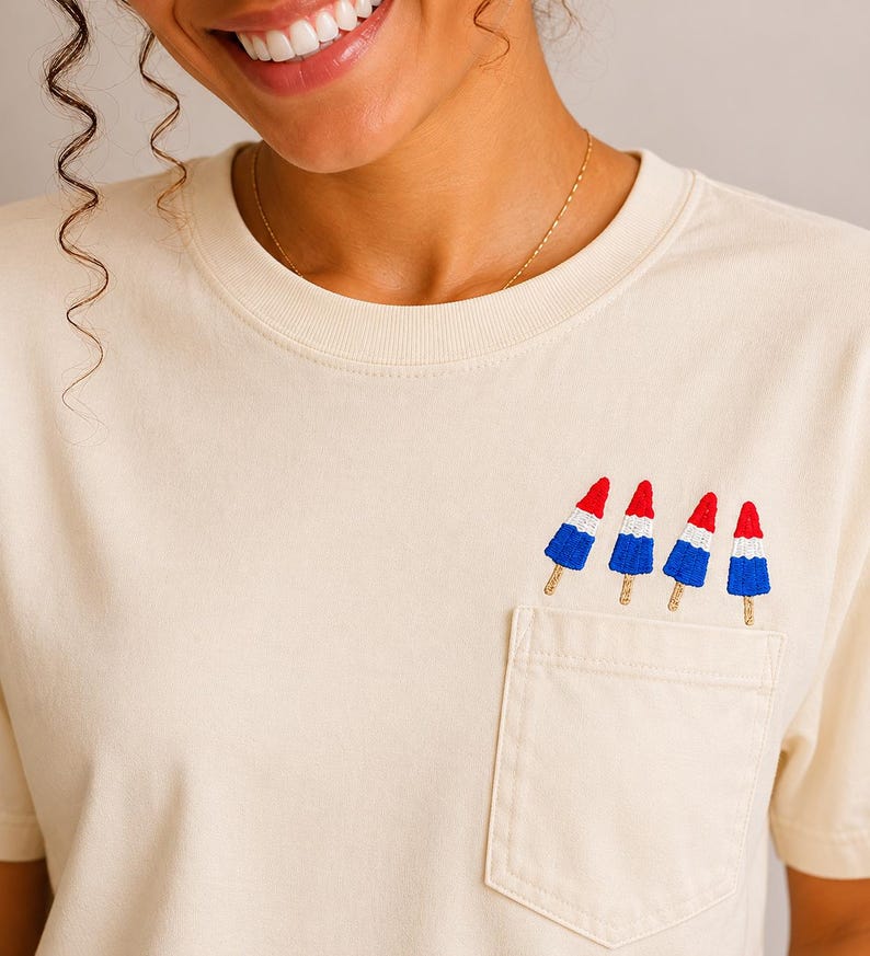 Op de afbeelding: Een cr&egrave;mekleurig T-shirt met korte mouwen en een borstzak. Boven de zak zijn vier kleine ijsjes geborduurd, elk met een rode bovenkant, een witte middenkant en een blauwe onderkant, afgewerkt met een lichtbruin houten stokje.