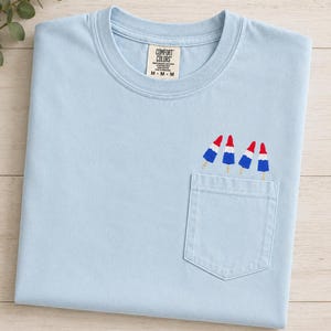 Op de afbeelding: Een lichtblauw T-shirt met korte mouwen, een ronde hals en een borstzak. Op de zak is een borduursel van vier rood-wit-blauwe ijsjes te zien. Het label in de nek bevat de tekst Comfort Colors M-M-M.