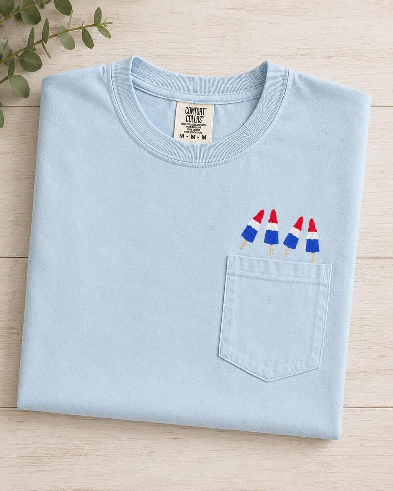 Op de afbeelding: Een lichtblauw T-shirt met korte mouwen, een ronde hals en een borstzak. Op de zak is een borduursel van vier rood-wit-blauwe ijsjes te zien. Het label in de nek bevat de tekst Comfort Colors M-M-M.