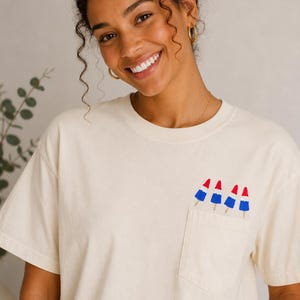 Op de afbeelding: Een cr&egrave;mekleurig T-shirt met korte mouwen en een borstzak. De zak is versierd met een grafische print van vier rood, wit en blauwe raketvormige ijsjes die in een rij langs de bovenrand van de zak zijn geplaatst.