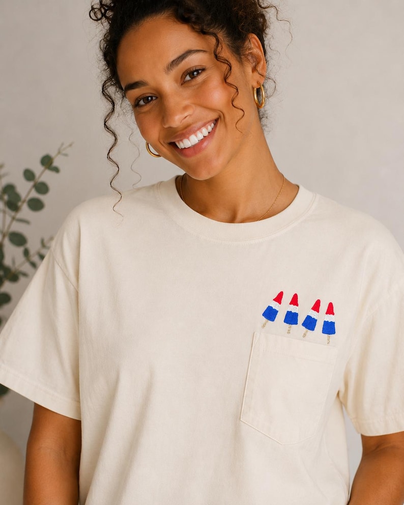 Op de afbeelding: Een cr&egrave;mekleurig T-shirt met korte mouwen en een borstzak. De zak is versierd met een grafische print van vier rood, wit en blauwe raketvormige ijsjes die in een rij langs de bovenrand van de zak zijn geplaatst.