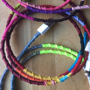 May include: A close-up of a colorful phone charging cable wrapped in yarn. The cable is wrapped in different colors, including red, orange, yellow, green, blue, purple, and pink. The cable is coiled up and the end of the cable is plugged into a phone charger.
