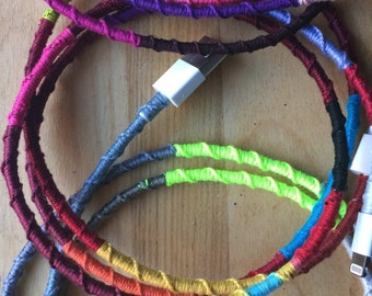 May include: A close-up of a colorful phone charging cable wrapped in yarn. The cable is wrapped in different colors, including red, orange, yellow, green, blue, purple, and pink. The cable is coiled up and the end of the cable is plugged into a phone charger.
