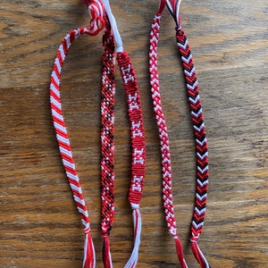 May include: Four friendship bracelets made with red, white, and black embroidery floss. The bracelets feature different patterns, including stripes, chevron, and a pixelated design.