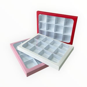 May include: Three candy boxes with clear lids. One box is red, another is white, and the third is pink. Each box has a grid of twelve square compartments. The boxes are empty and ready to be filled with treats.