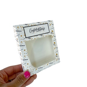 May include: White gift box with a window, decorated with gold stars, graduation caps, and the words "Congratulations!" in black lettering.