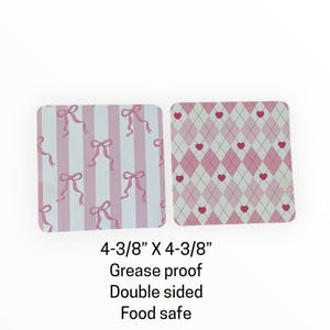 May include: Two square, double-sided, food-safe coasters. One has a light blue and pink striped pattern with pink bows. The other has a pink and white argyle pattern with pink hearts. Each coaster measures 4-3/8" x 4-3/8".