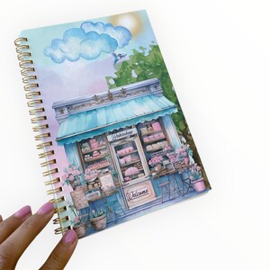 May include: Spiral-bound notebook featuring a watercolor illustration of a bakery with a blue awning and pink flowers. The word "Bakeshop" is above the display window, and "Welcome" is on the door. A blue bird flies near a cloud and sun.