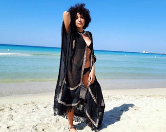 May include: A woman wearing a black sheer beach cover-up with fringe trim and a black bikini top. She is standing on a sandy beach with the sea in the background.