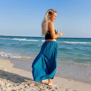 May include: A woman wearing a long, flowing turquoise skirt stands on a sandy beach with the ocean in the background.