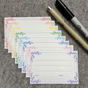 May include: A stack of white note cards with colorful floral borders and lined interiors. The borders feature a rainbow of colors, and the cards have the words "Love" and "Date" printed on them. Two pens, one black and one beige, are next to the cards.