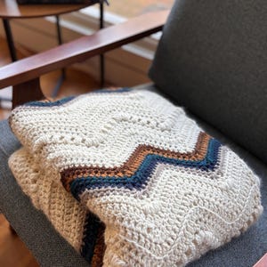 Puede incluir: Una manta de ganchillo plegada, de color crema, con un patrón de chevron en marrón y verde azulado. La manta está sobre el brazo de una silla gris. La textura de la manta es visible, con un patrón de puntadas detallado.