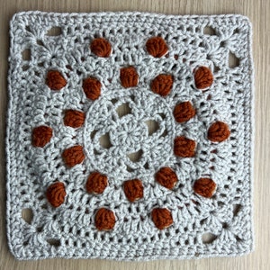 Op de afbeelding: Een vierkant gehaakt granny square met een cirkelvormig patroon van bruine garenknopjes in het midden. Het vierkant is gemaakt met lichtgrijs garen.