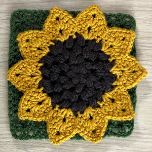 May include: A crocheted sunflower with a dark brown center and yellow petals. The sunflower is set against a green crocheted square.