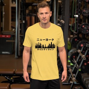 May include: A yellow t-shirt featuring a black silhouette of the New York City skyline, including the Statue of Liberty. The Japanese text above the skyline reads "New York" and the text below reads "My City Life".