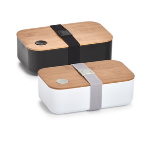 Könnte beinhalten: Zwei Lunchboxen mit Bambusdeckeln und einem Riemenverschluss. Eine Lunchbox ist schwarz und die andere ist weiß.