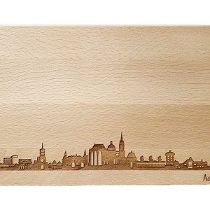 Könnte beinhalten: Ein Holzbrett mit einem lasergravierten Skyline von Aachen, Deutschland. Die Gebäude der Stadt sind in Silhouette dargestellt, mit dem Text "Aachen" unten rechts.