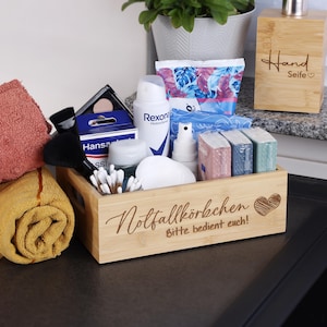 Könnte beinhalten: Ein Bambus-Organizer gefüllt mit Toilettenartikeln, darunter Flaschen, Wattestäbchen und Make-up. Der Organizer hat ein Herz und den Text "Notfallkörbchen Bitte bedient euch!". Ein Seifenspender aus Holz und gerollte Handtücher sind ebenfalls sichtbar.