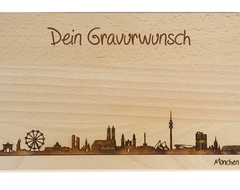 May include: A wooden cutting board with a laser-engraved skyline of Munich, Germany. The board has the text "Dein Gravurwunsch" at the top and "München" at the bottom.