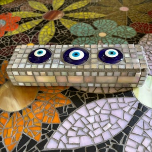 May include: A small, rectangular, gray mosaic tile with three blue evil eye charms. The tile is made of small, white and gray mosaic tiles.