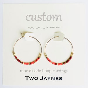 K&ouml;nnte beinhalten: Goldene Creolen mit roten und orangen Perlen. Die Ohrringe befinden sich auf einer wei&szlig;en Karte mit dem Text "custom" und "morse code hoop earrings" sowie dem Namen "TWO JAYNES".