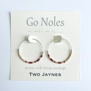 May include: A pair of silver hoop earrings with maroon and beige beads. The earrings are displayed on a card with the text "Go Noles" and "morse code hoop earrings" and the brand name "TWO JAYNES".