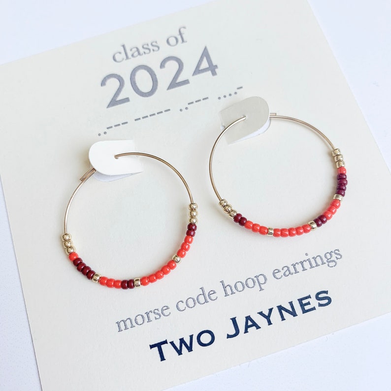 K&ouml;nnte beinhalten: Goldene Creolen mit roten und kastanienbraunen Perlen. Die Ohrringe werden auf einer Karte pr&auml;sentiert, auf der "Klasse von 2024" und "Morse-Code-Creolen" sowie der Markenname "Two Jaynes" stehen.