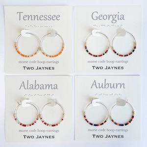 K&ouml;nnte beinhalten: Vier Karten mit verschiedenen College-Teamnamen und -farben. Jede Karte zeigt ein Paar silberne Creolen mit bunten Perlen. Die Karten lauten "Tennessee", "Georgia", "Alabama" und "Auburn". Jede Karte enth&auml;lt auch den Text "morse code hoop earrings" und "TWO JAYNES".