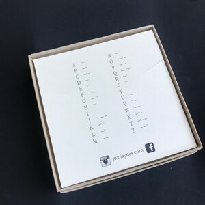K&ouml;nnte beinhalten: Eine wei&szlig;e Karte mit dem Alphabet in Morsecode. Die Karte befindet sich in einer braunen Schachtel mit einem Social-Media-Symbol und der Website twojaynes.com.