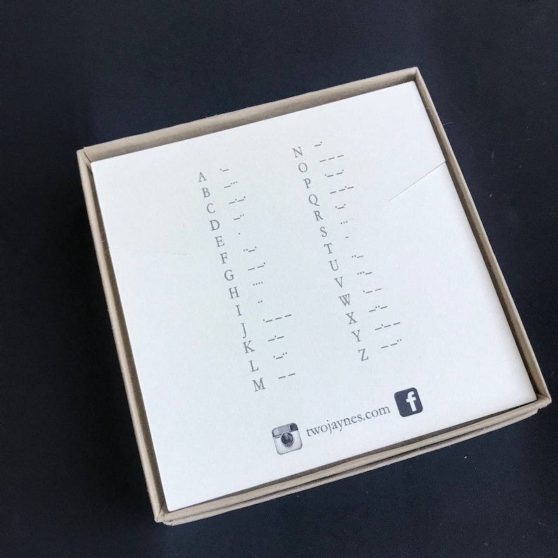 K&ouml;nnte beinhalten: Eine wei&szlig;e Karte mit dem Alphabet in Morsecode. Die Karte befindet sich in einer braunen Schachtel mit einem Social-Media-Symbol und der Website twojaynes.com.