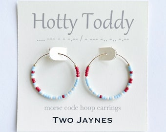 May include: A pair of silver hoop earrings with white, blue, and red beads. The earrings are displayed on a white card with the text "Hotty Toddy" and "Morse code hoop earrings" and the brand name "TWO JAYNES".