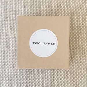 K&ouml;nnte beinhalten: Ein brauner Karton mit einem wei&szlig;en runden Aufkleber, auf dem in schwarzer Schrift "TWO JAYNES" steht.