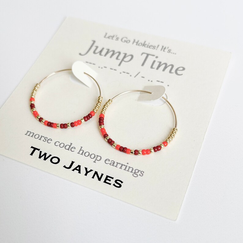 K&ouml;nnte beinhalten: Zwei goldene Creolen mit roten und orangefarbenen Perlen. Die Ohrringe befinden sich auf einer wei&szlig;en Karte mit dem Text "Let's Go Hokies! It's... Jump Time" und "morse code hoop earrings TWO JAYNES".