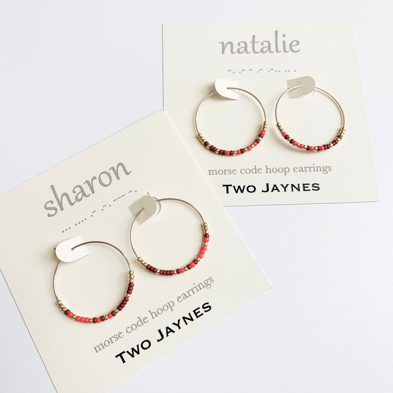 K&ouml;nnte beinhalten: Zwei Paar goldene Creolen mit roten und goldenen Perlen. Die Ohrringe werden auf wei&szlig;en Karten mit den Namen "Natalie" und "Sharon" und dem Text "Morsecode-Creolen TWO JAYNES" pr&auml;sentiert.