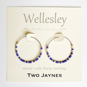 May include: A pair of gold hoop earrings with blue and white beads. The earrings are displayed on a white card with the text "Wellesley" and "morse code hoop earrings" and the logo "TWO JAYNES".